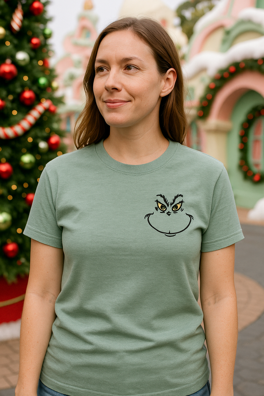 Person wearing a green t-shirt with a smirking grinch face design in front of a decorated Christmas tree.
